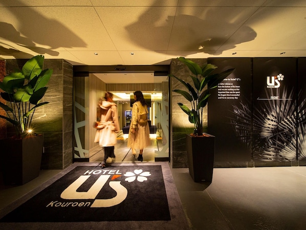 A hotel entrance with a black carpet featuring the hotel logo and name. There are large potted plants on either side of the entrance.