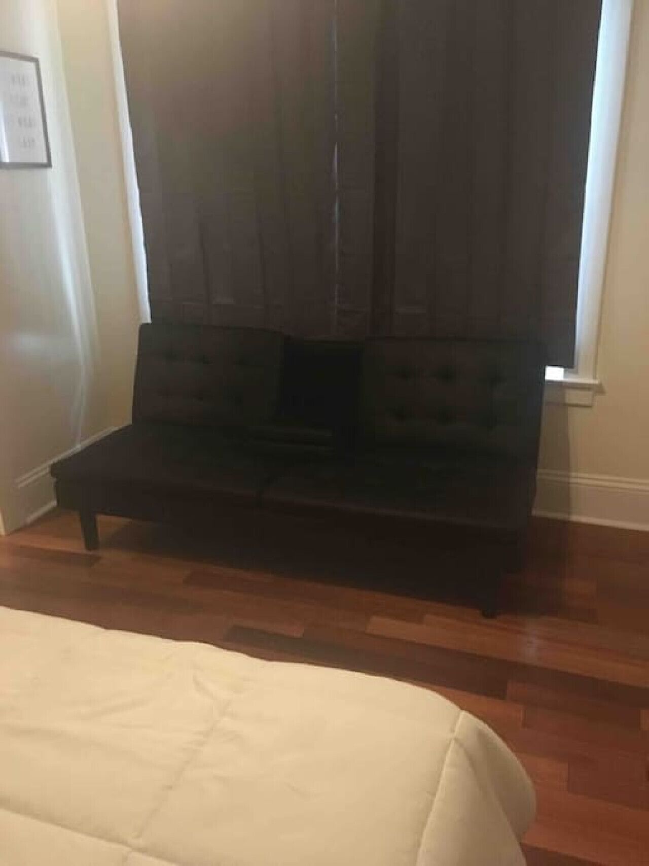 Une pièce avec un canapé de couleur sombre placé contre un mur, un lit blanc avec une courtepointe et un plancher en bois.