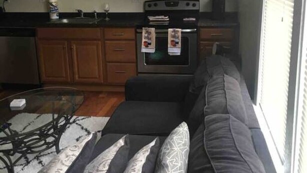 A living room with a gray sofa, a glass coffee table, and a kitchen area with wooden cabinets, a microwave, and an oven. - The couples home with free wifi (Schenectady)