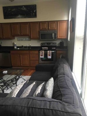 Una sala de estar con un sofá gris, una mesa de centro de vidrio y una zona de cocina con armarios de madera, un microondas y un horno.