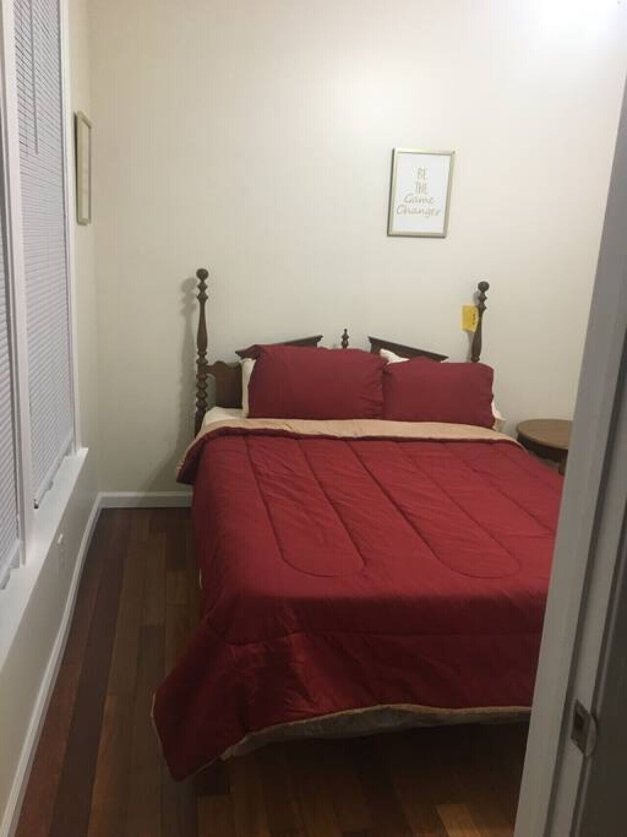 A small bedroom with a bed, a nightstand, a framed quote on the wall, and a window with blinds.