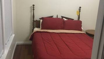 A small bedroom with a bed, a nightstand, a framed quote on the wall, and a window with blinds.