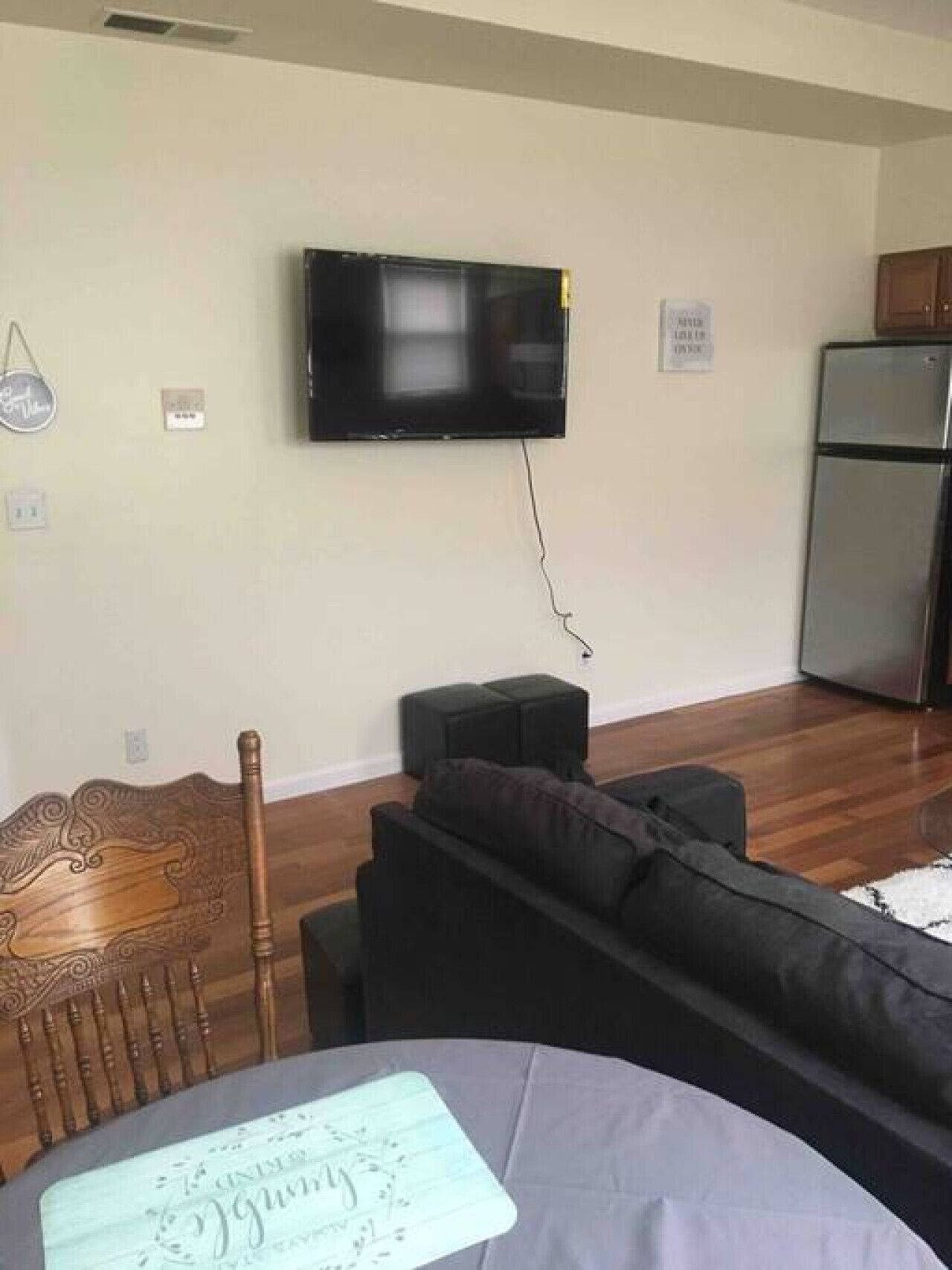 Una sala de estar con una televisión de pantalla plana montada en la pared, una mesa de comedor de madera con sillas, un sofá negro y un refrigerador al fondo.