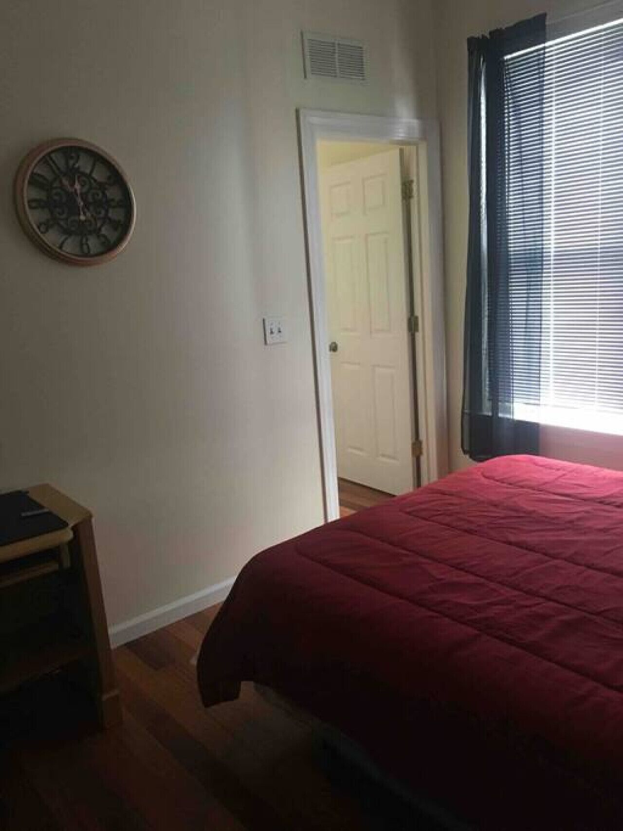 Un dormitorio con una colcha roja, una mesita de noche de madera, un reloj de pared y una puerta que da a otra habitación.