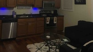 A compact kitchen with wooden cabinets, a microwave, and a dishwasher. There's a ceiling fan, a painting on the wall, and a small table with a glass top. - The couples home with free wifi (Schenectady)