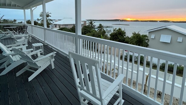 En overdækket terrasse med hvide gyngestole og udsigt til solnedgang over vandet.