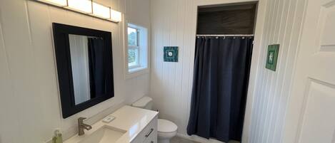 Et badeværelse med hvid kommode, spejl, toilet, bruseforhæng og et lille vindue.