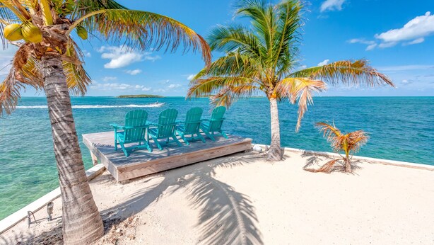A beach with a wooden deck, three turquoise chairs, and palm trees. - Waterfront 4-Bedroom Home on Private Peninsula w/ Private Dock! (Elbow Cay)