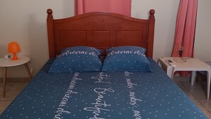 Un dormitorio con una cama de madera, ropa de cama azul con texto blanco, una mesita de noche pequeña con una lámpara y una ventana con cortinas rosadas.