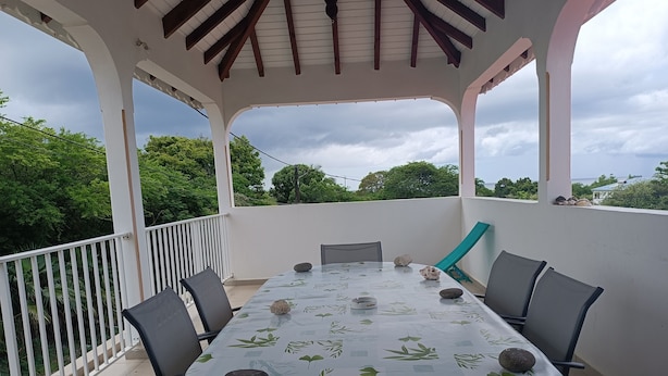Un área de comedor al aire libre con techo, con una mesa preparada para cuatro personas, rodeada de sillas y una vista de follaje y árboles al fondo.