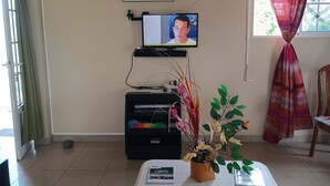 Una sala de estar con televisor, un cuadro enmarcado, una mesita con revistas y una planta en maceta.