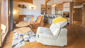 Amplia sala de estar con un sofá, sillones y una mesa de centro. Se ve una cocina con electrodomésticos modernos al fondo.