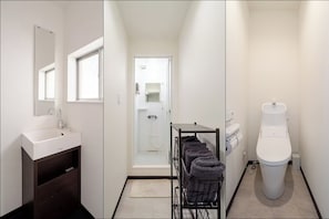 Un bany amb un lavabo blanc, mirall i vàter, una zona de dutxa i un prestatge amb tovalloles.