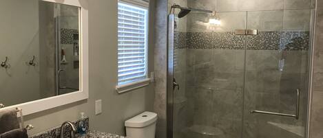 A bathroom with a granite countertop sink, a toilet, a glass-enclosed shower, and a window with blinds.