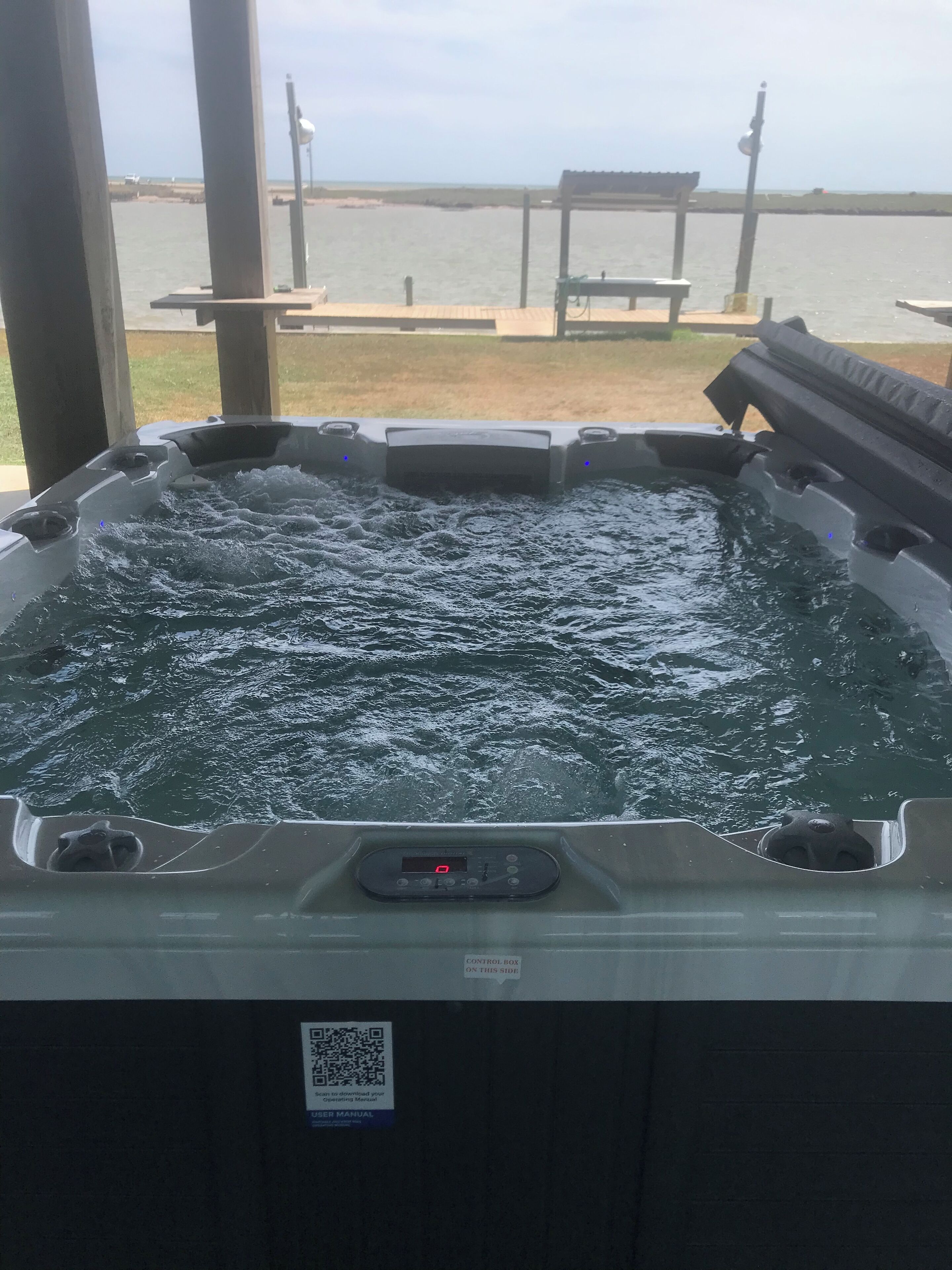 Un jacuzzi con panel de control y un código QR, rodeado de pilares de madera, con vistas a un cuerpo de agua y una banca al fondo.