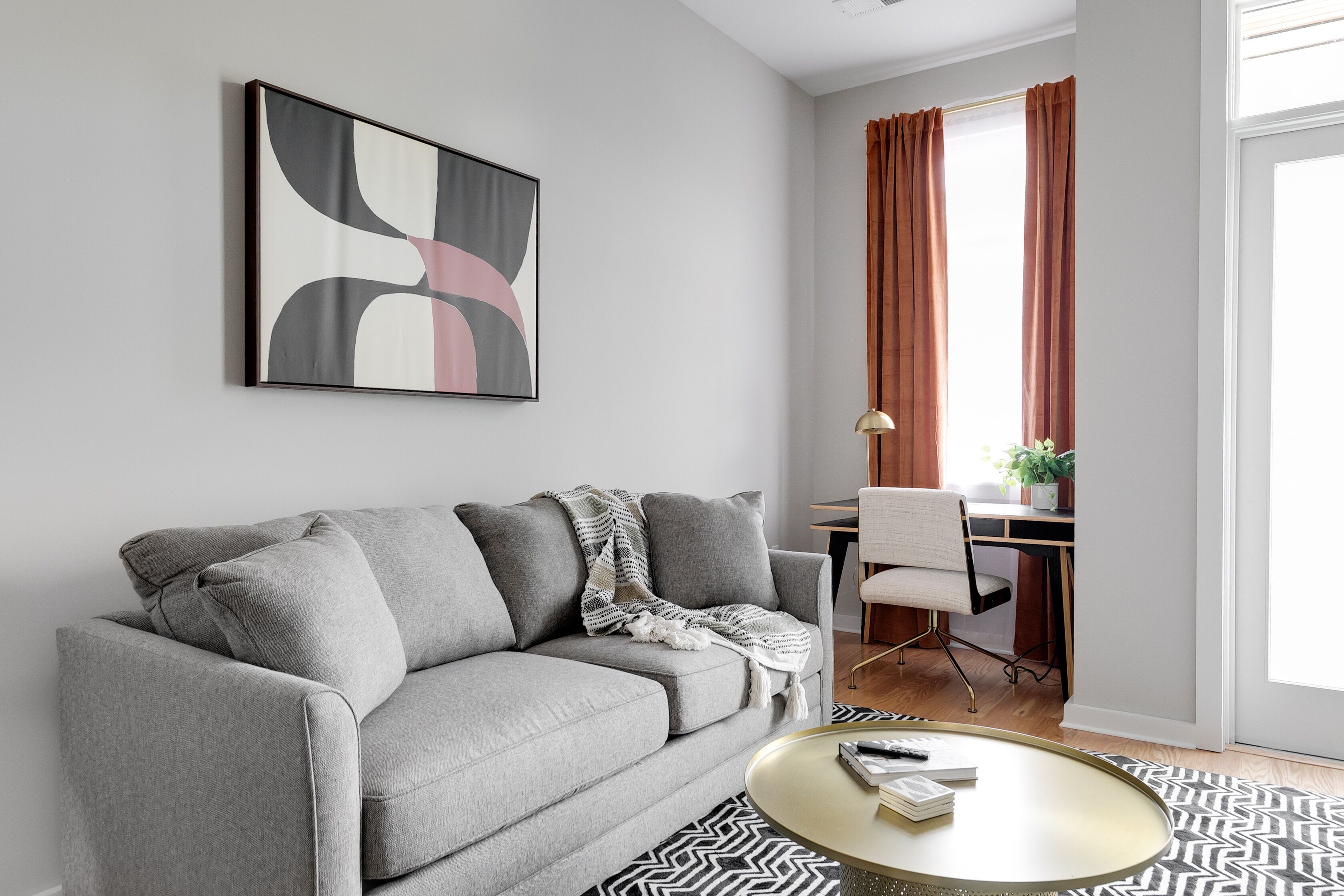 Un salon moderne avec un canapé gris, une table basse ronde et un tapis noir et blanc. On aperçoit un bureau avec une chaise et une plante au fond.