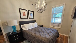 Ein Schlafzimmer mit einem Bett, Nachttischen, einer Lampe, einer Chandelier, einem Fenster mit Jalousien und einem gemusterten Teppich.