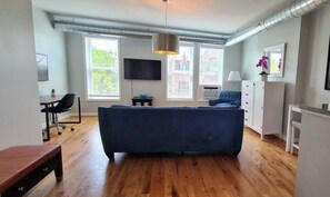 A living room with a blue sofa, wooden flooring, a TV mounted on the wall, a desk with a chair, and a window with a view of buildings.