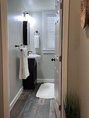 Un couloir étroit mène à une salle de bain équipée d’un lavabo blanc, d’un miroir et d’un porte-serviettes.
