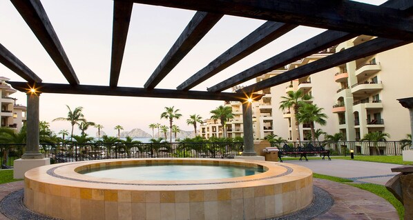 Un bain à remous circulaire, installé sous une pergola en bois, offre une vue sur un bâtiment d’un complexe hôtelier et sur des palmiers.