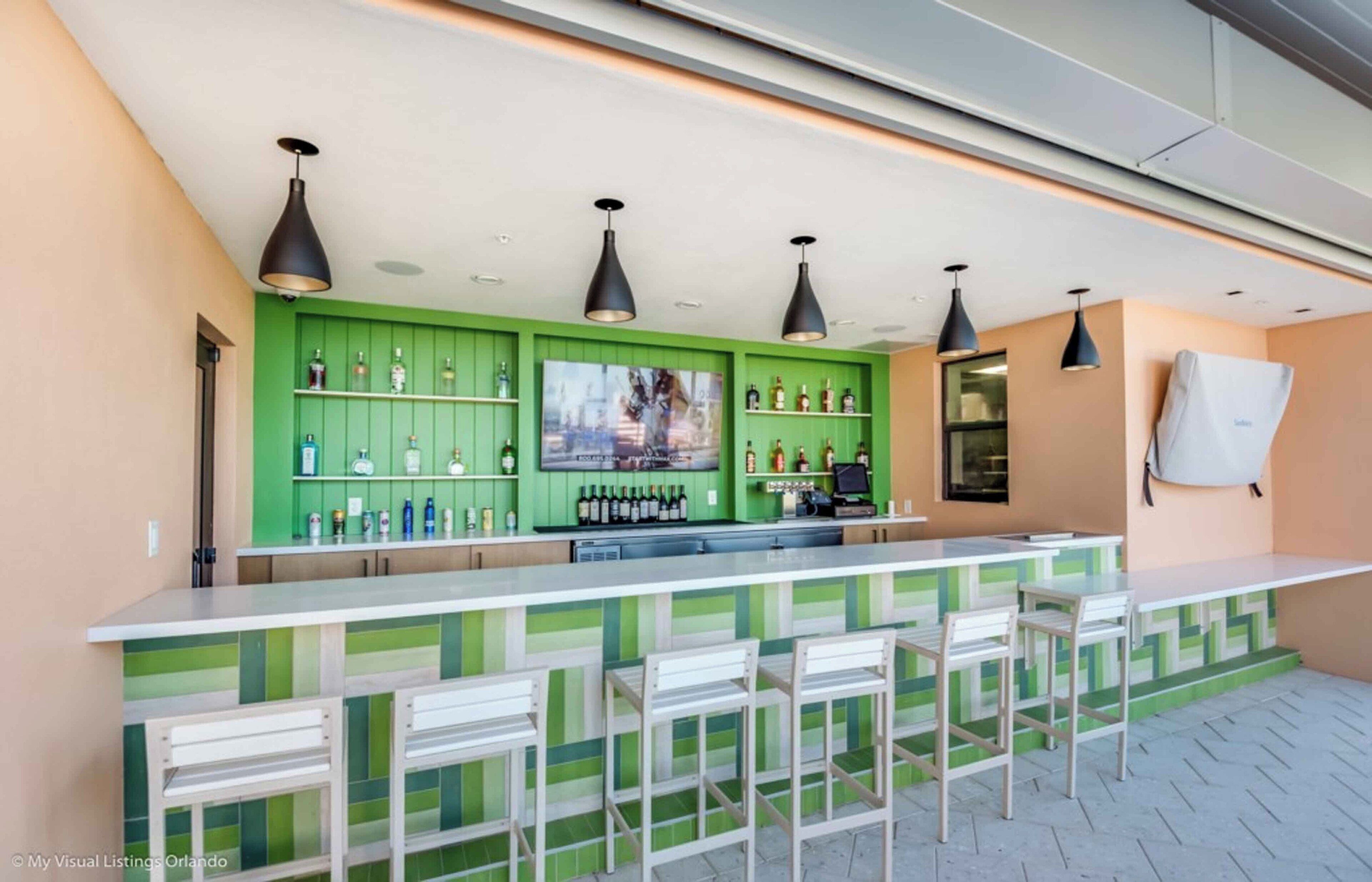 Een bar met groene toogbladen en witte stoelen. Er staan meerdere flessen en een televisiescherm op de bar.