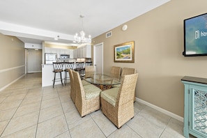 A dining area with a glass table and wicker chairs, a kitchen in the background, and a teal cabinet with a decorative design.