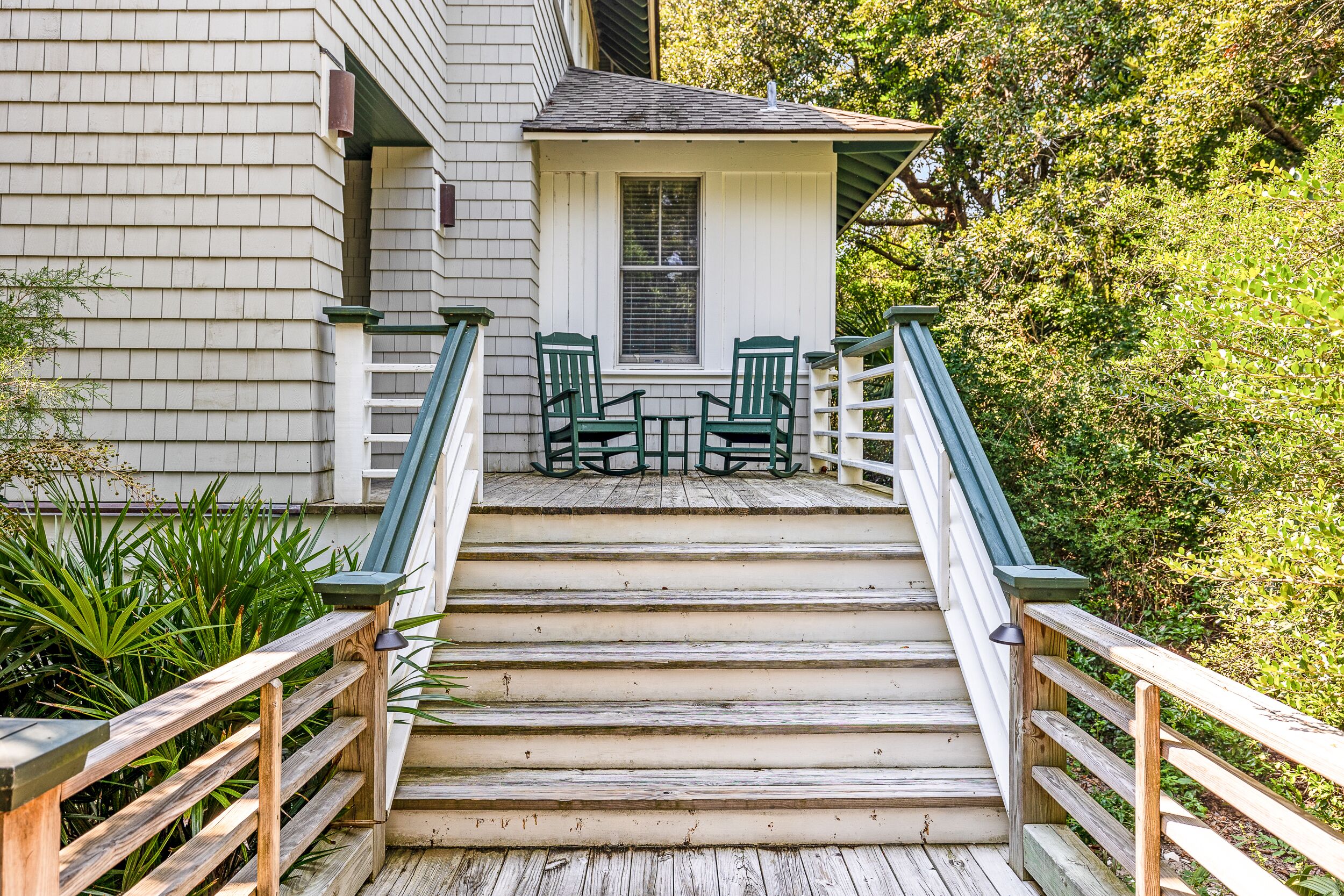 Un escalier en bois mène à une véranda où se trouvent des chaises vertes et une petite table.