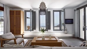 A modern living room with a sofa, a coffee table, and a view of the ocean through large windows. - Villa Angeliki - Private villa with pool in Santorini (Thera)
