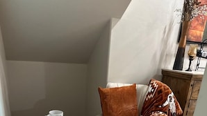 Een gezellige hoek met een witte fauteuil, een decoratief kussen, een klein rond tafeltje met kop en schotel, en een witte poef.