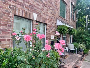 A brick building with a number '5208' on the window, a rose bush in the foreground, and a chair and potted plant on the sidewalk.