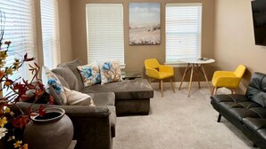 Een woonkamer met een hoekbank, een zwarte leren bank, een gele fauteuil en een klein rond tafeltje.