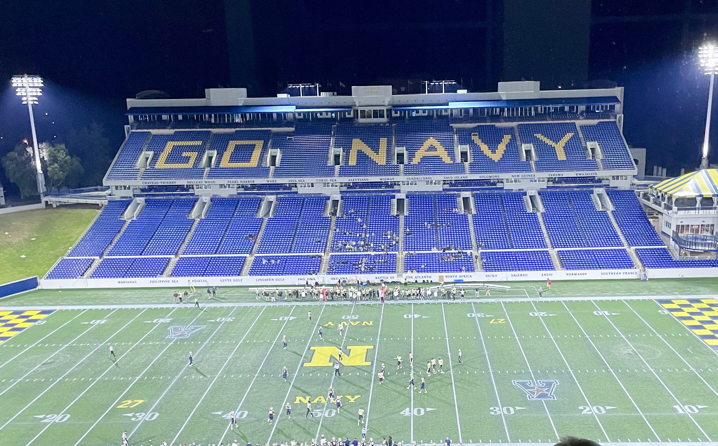 A football stadium with blue seating and a field marked with yard lines and team logos.