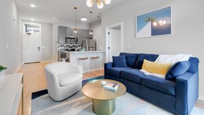 Ein modernes Wohnzimmer mit einem blauen Sofa, einem weißen Sessel und einem runden Couchtisch. Im Hintergrund ist eine Küche mit weißen Schränken und einer Mikrowelle erkennbar.