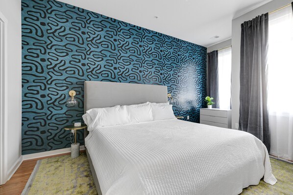 Quarto com parede estampada, cama com roupa de cama branca, mesa de cabeceira com abajur e janela com cortinas.