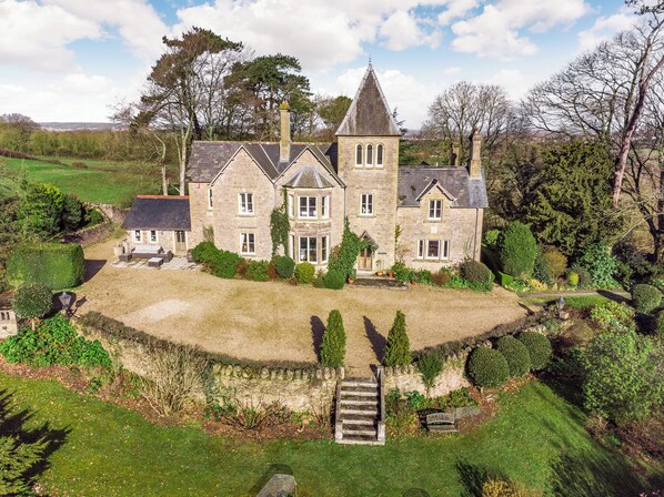 A large stone house with multiple windows, a central tower, and a well-maintained garden.