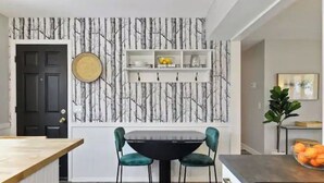 Un comedor con una mesa negra y sillas color esmeralda, una puerta negra y una pared con diseño de árbol.