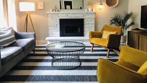Una sala de estar con chimenea, un sofá, un sillón amarillo, una mesa de centro y un tapete a rayas.
