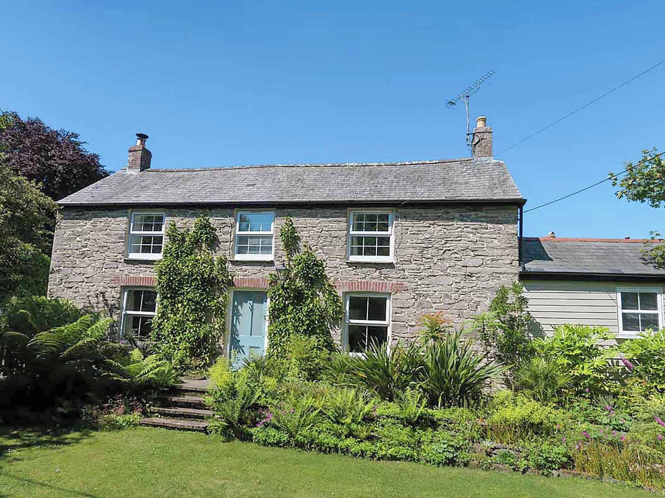 Une maison en pierre à deux étages, avec une porte bleue, entourée d’une végétation luxuriante et d’une pelouse bien entretenue.