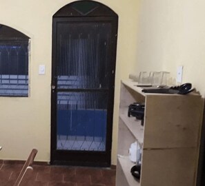 A room with a tiled floor, a black door with a metal grill, a window with a metal grill, and a shelf with various items. - Sana Aluguel Economico (Macaé)