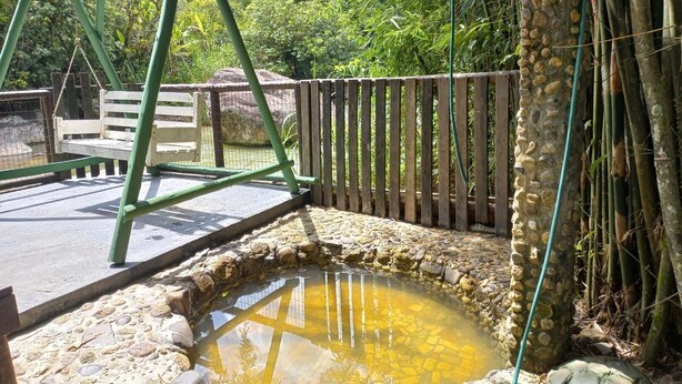 A stone-walled pond with a wooden swing set and a fence in a natural setting. - Sana Aluguel Economico (Macaé)