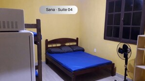 A small, single-bed room with a wooden headboard, a blue mattress, and a standing fan. - Sana Aluguel Economico (Macaé)