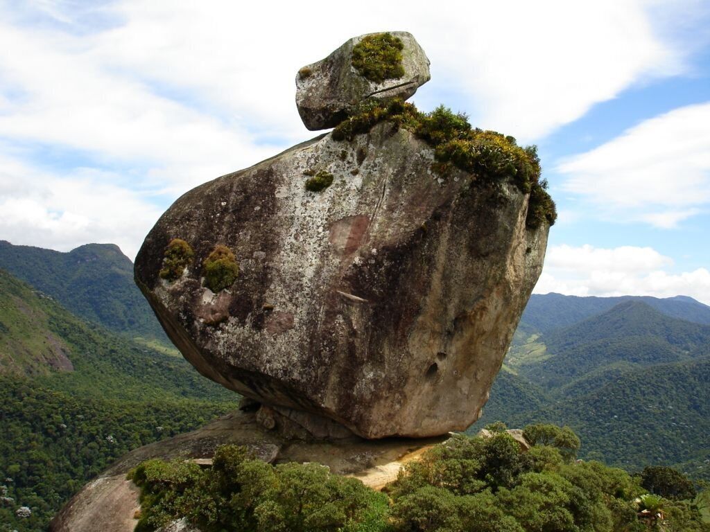 Una gran formación rocosa con una roca equilibrada en la cima, rodeada de exuberantes montañas verdes y un cielo despejado.