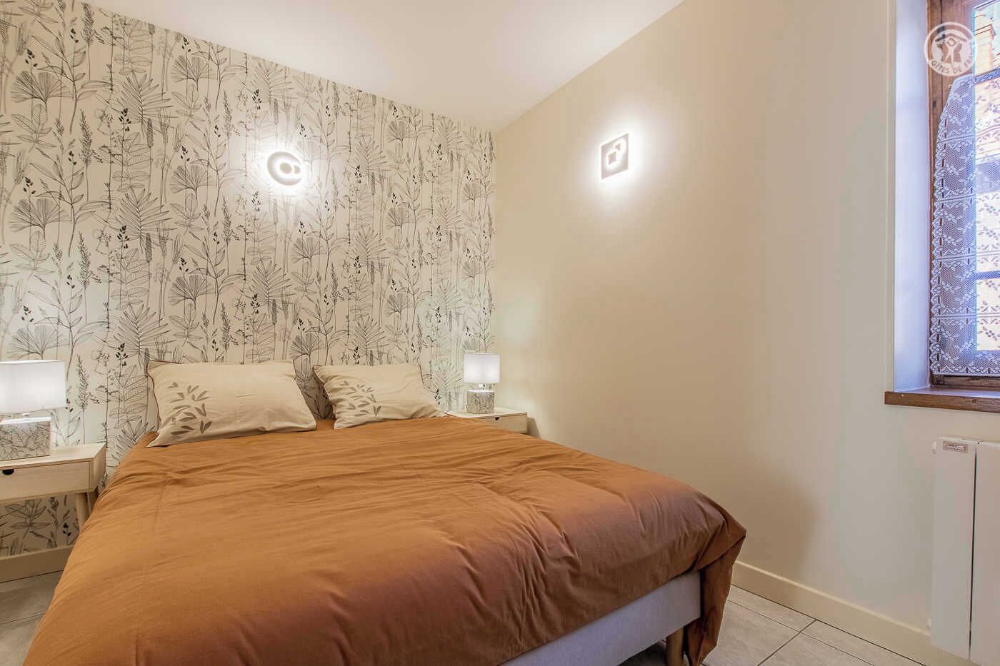 Un dormitorio con papel tapiz floral, un edredón marrón, dos mesitas de noche con lámparas y una ventana con cortinas de encaje.