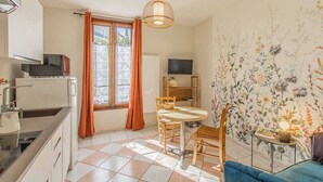 A compact kitchen with a refrigerator, microwave, and stove. There is a dining area with a table and chairs, and a television on a stand. The room has a window with curtains and a floral wallpaper.