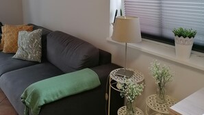 A cozy living room with a dark grey sofa, a green throw blanket, and a white lamp on a small table. There are two white decorative vases on a shelf by the window.