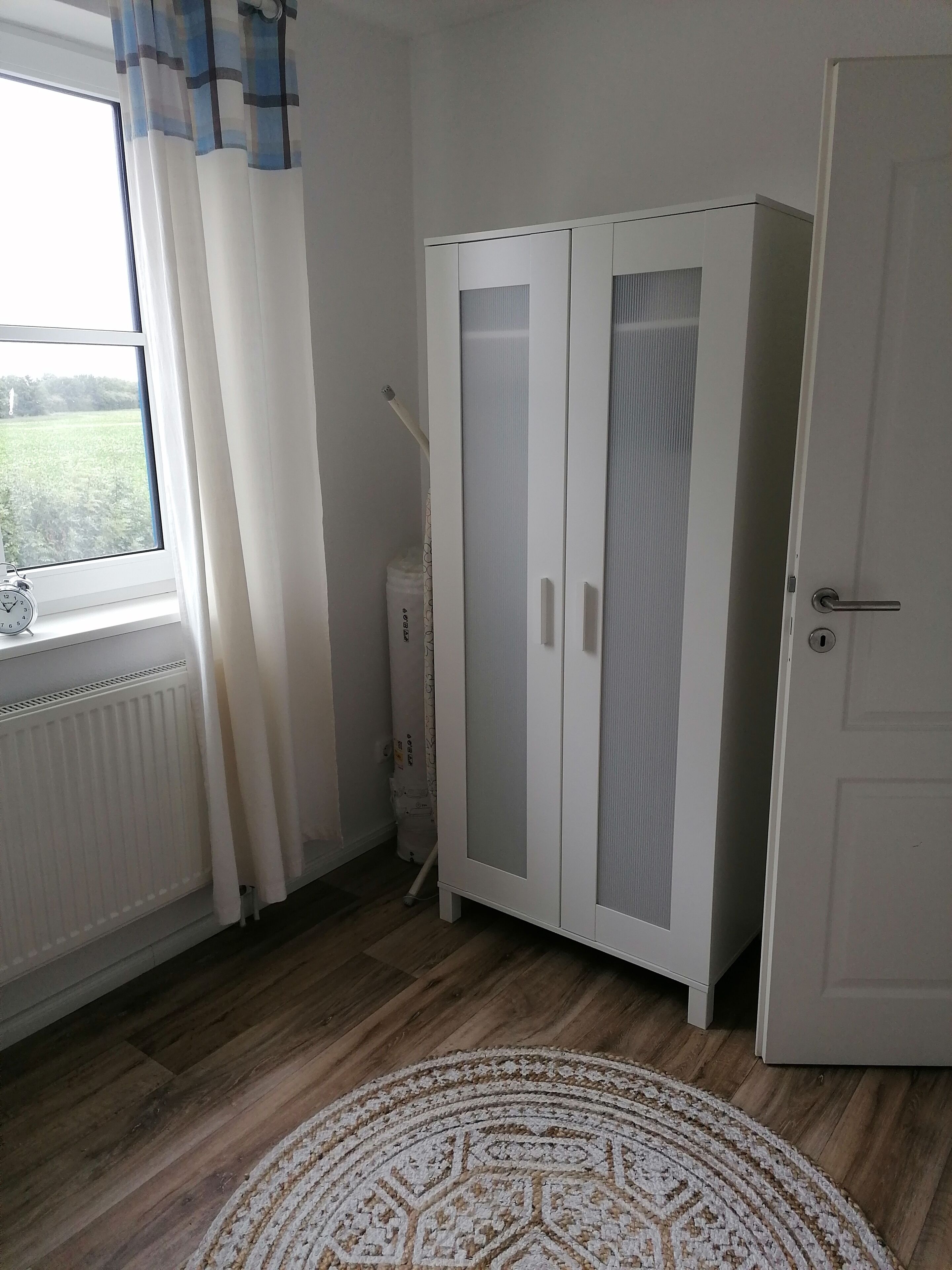 Une pièce avec une armoire blanche, un radiateur, une fenêtre avec des rideaux et un tapis à motifs.