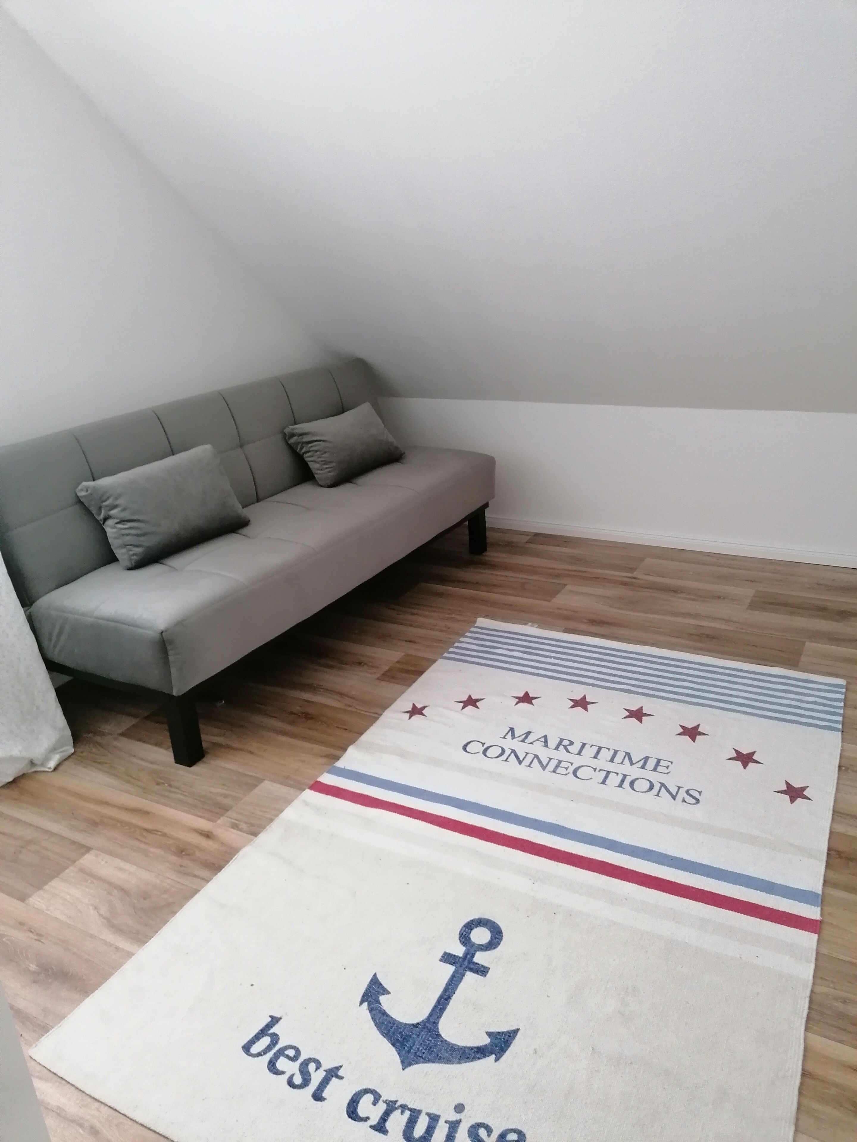 Ein Zimmer mit grauem Sofa, Holzboden und Teppich mit maritimen Motiven und Anker.