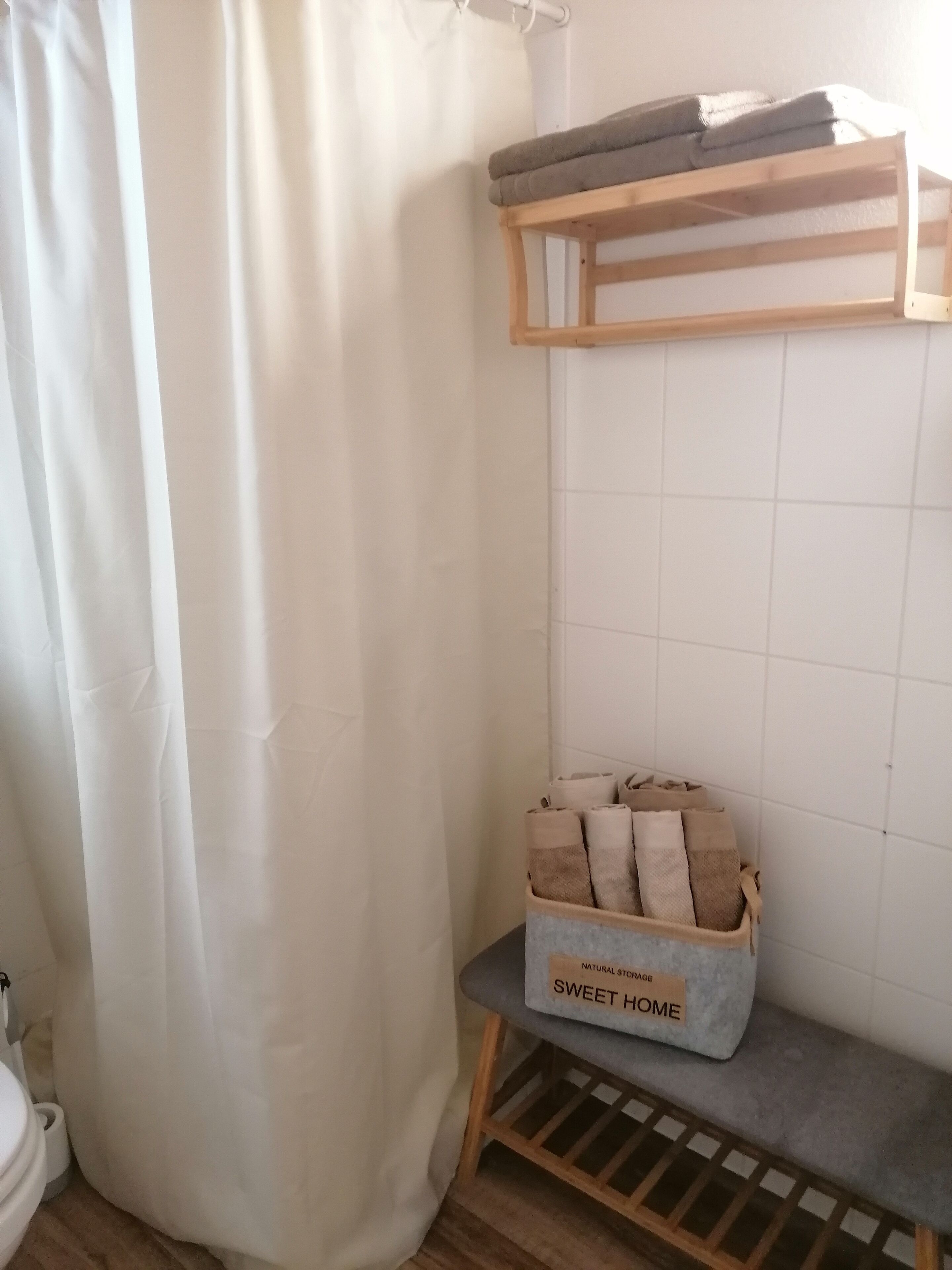 A bathroom with a shower curtain, a wooden shelf with towels, and a storage box labeled 'SWEET HOME' containing towels.