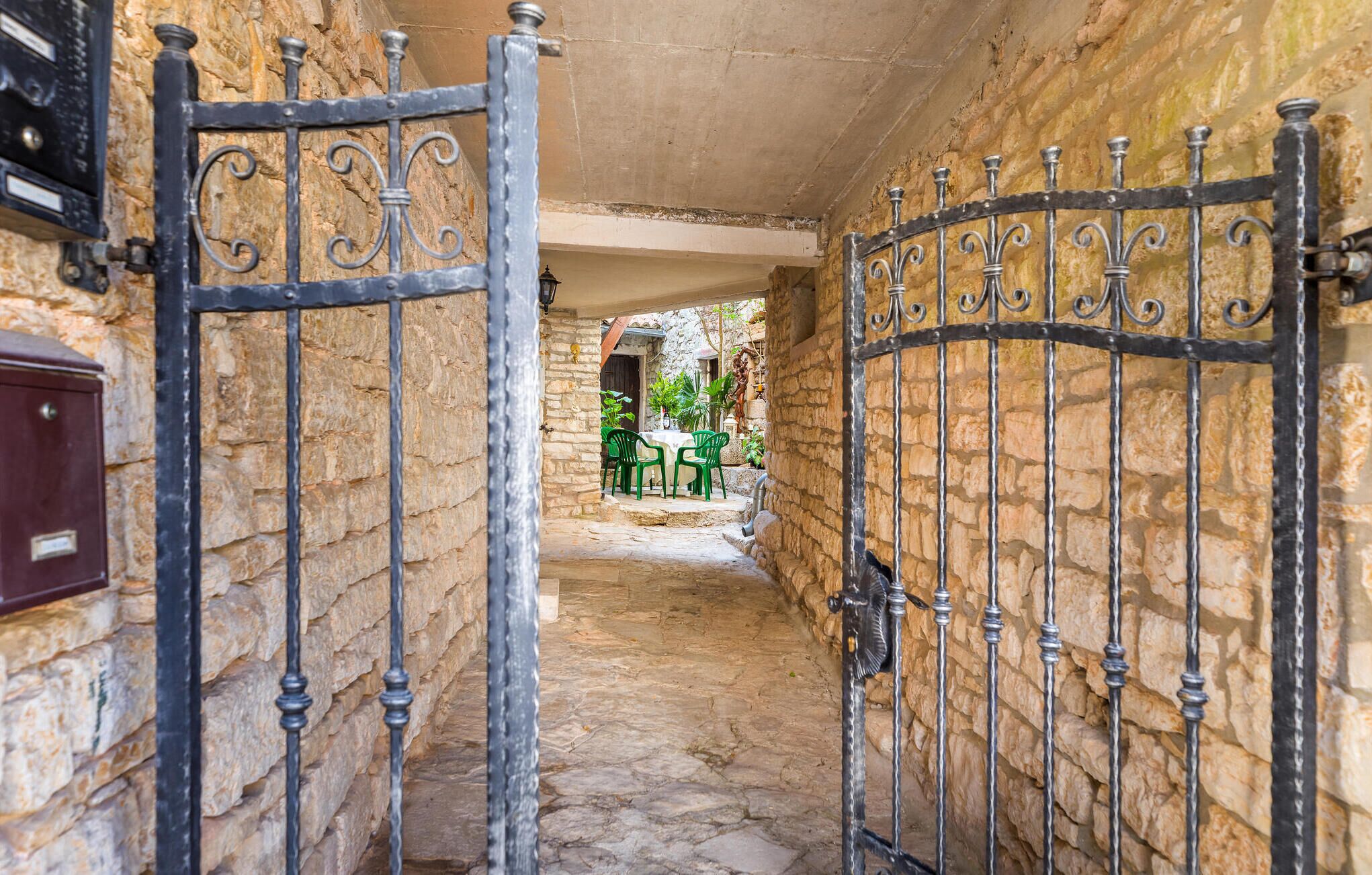 Un estrecho pasillo de piedra con una reja metálica, que conduce a un patio con sillas verdes y una mesa.
