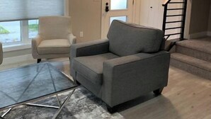 Ein Wohnzimmer mit grauem Sofa, einem weißen Sessel und einem Glastisch. Eine Treppe mit schwarzer Geländer führt zu einer Tür mit sandgestrahlten Glasfeldern.